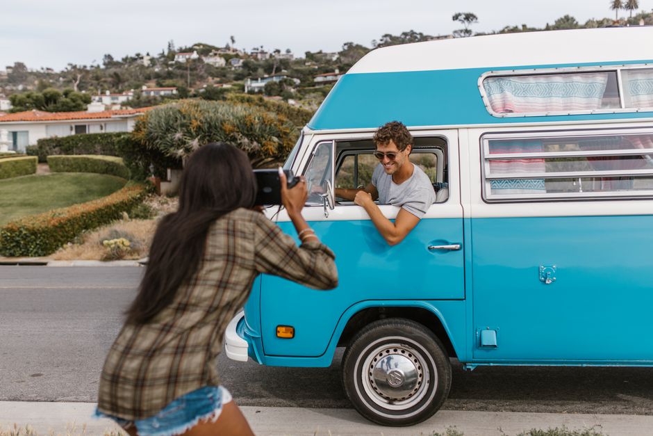 A young man with curly hair and wearing sunglasses, smiling and leaning out of the driver’s side window of a vintage blue camper van, while a young woman with long dark hair in a plaid shirt, denim 