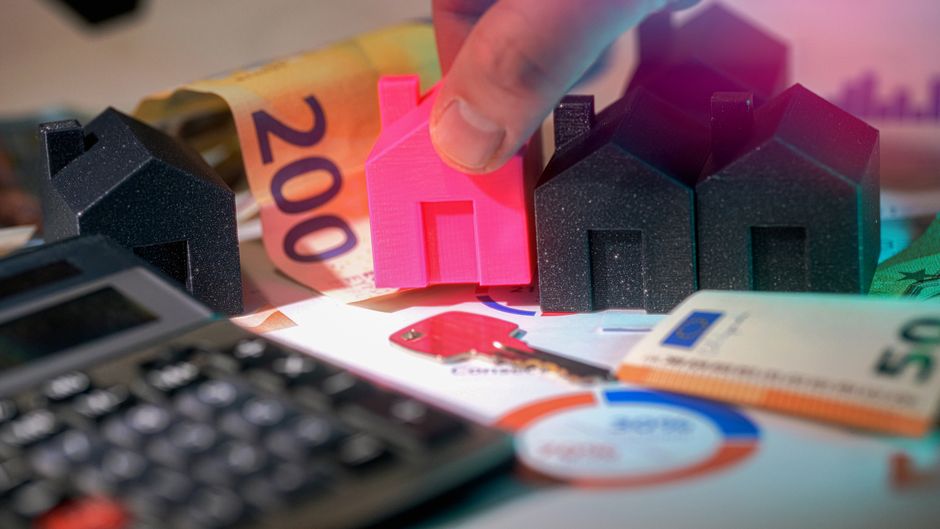 Close-up view of a person's hand holding a small pink house-shaped model, positioned among several black miniature house models on a cluttered surface. The area features various items including Euro b