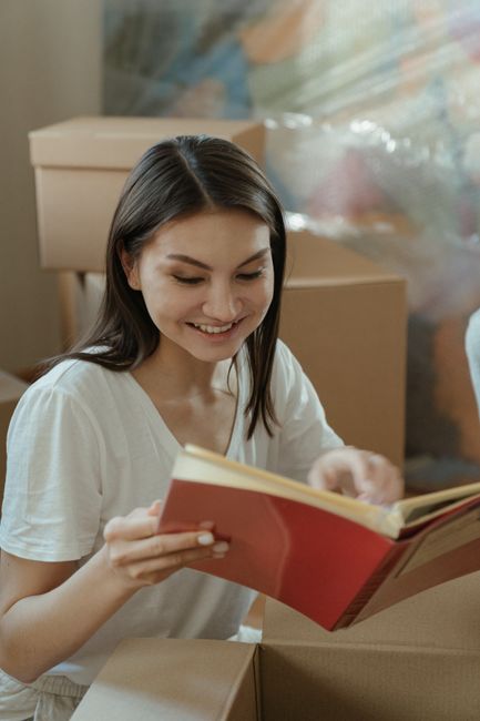 A young woman with long brown hair, wearing a white T-shirt, is inside a room surrounded by cardboard boxes. She is smiling while reading a book, with her head slightly tilted down towards the open pa